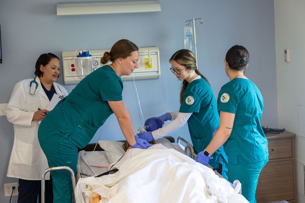 A healthcare team works together around a patient in a hospital bed. Three nurses in teal scrubs provide care while a medical professional in a white coat observes and takes notes. The patient is covered with a white blanket, with medical equipment and an IV visible in the background.