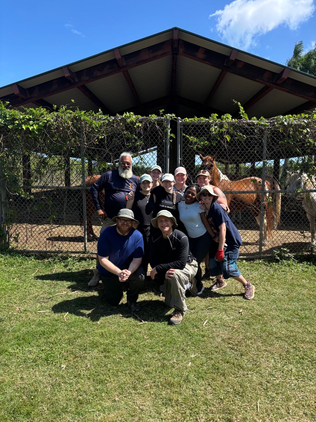 A volunteer group poses in front of a fenced horse enclosure on a bright sunny day. Several horses stand behind the group beneath a covered shelter, while the volunteers wear hats, outdoor clothing, and smiles during what appears to be a community or service activity.