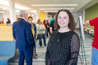 A young woman with curly hair smiles at the camera in a bright, modern indoor space, while small groups of people chat in the background near seating and a presentation easel.